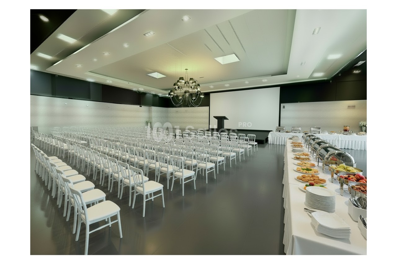 Salle de conférence avec rangées de chaises blanches, un podium, un écran de projection et un buffet dressé sur le côté.