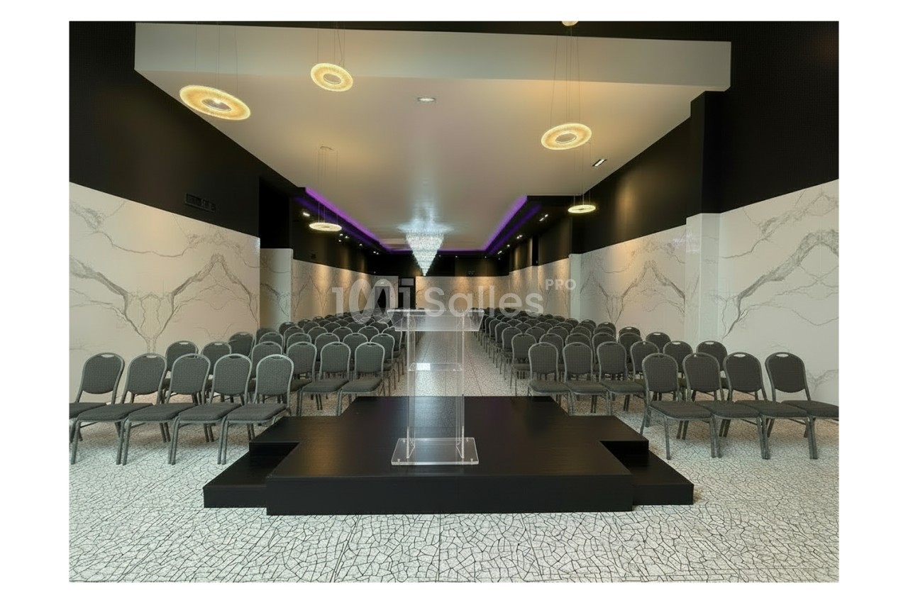 Salle de conférence moderne avec rangées de chaises, estrade centrale et éclairage suspendu élégant.