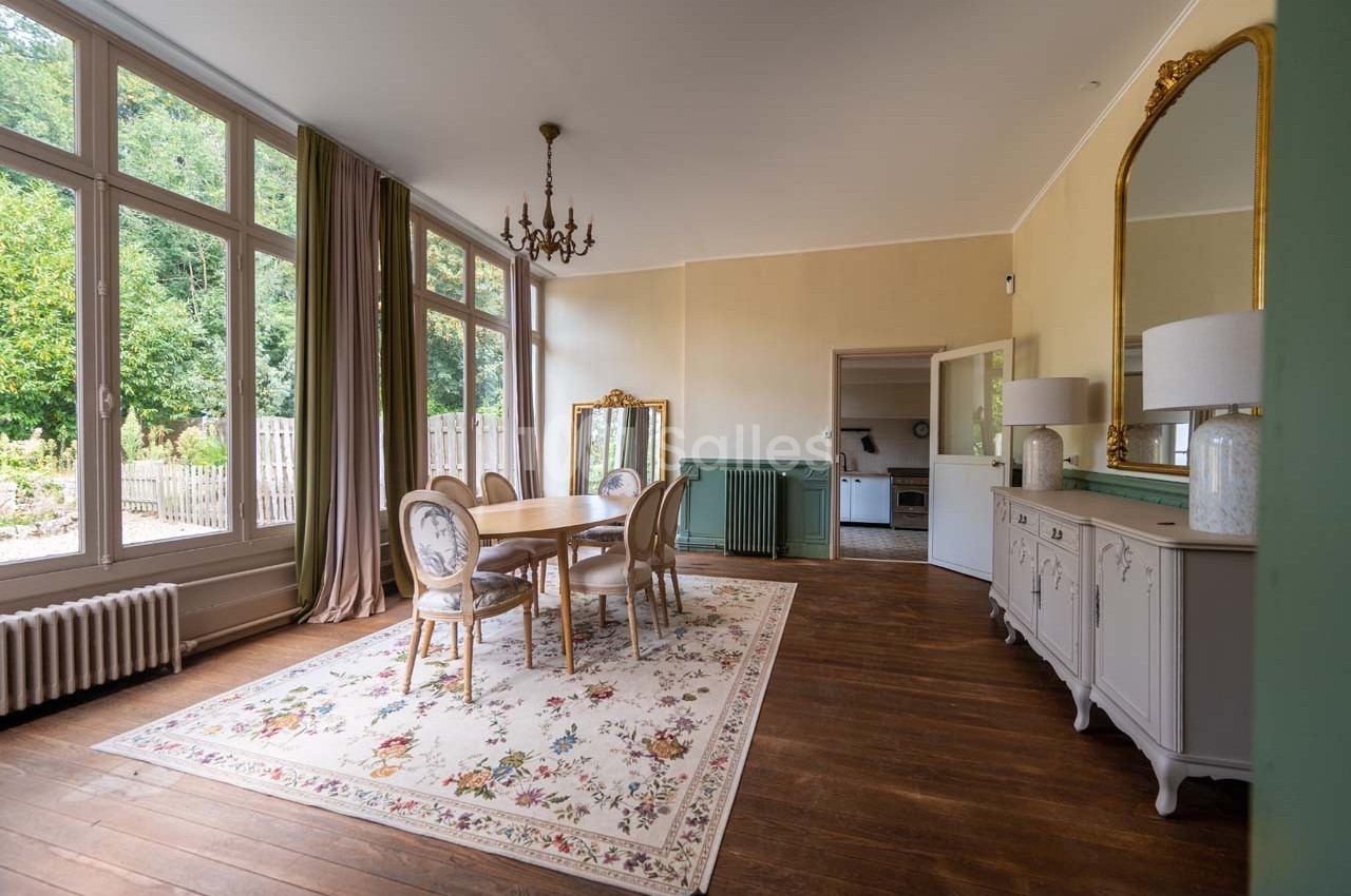 Salle à manger lumineuse avec table en bois, chaises élégantes, tapis fleuri, miroir doré et grandes fenêtres donnant sur un…