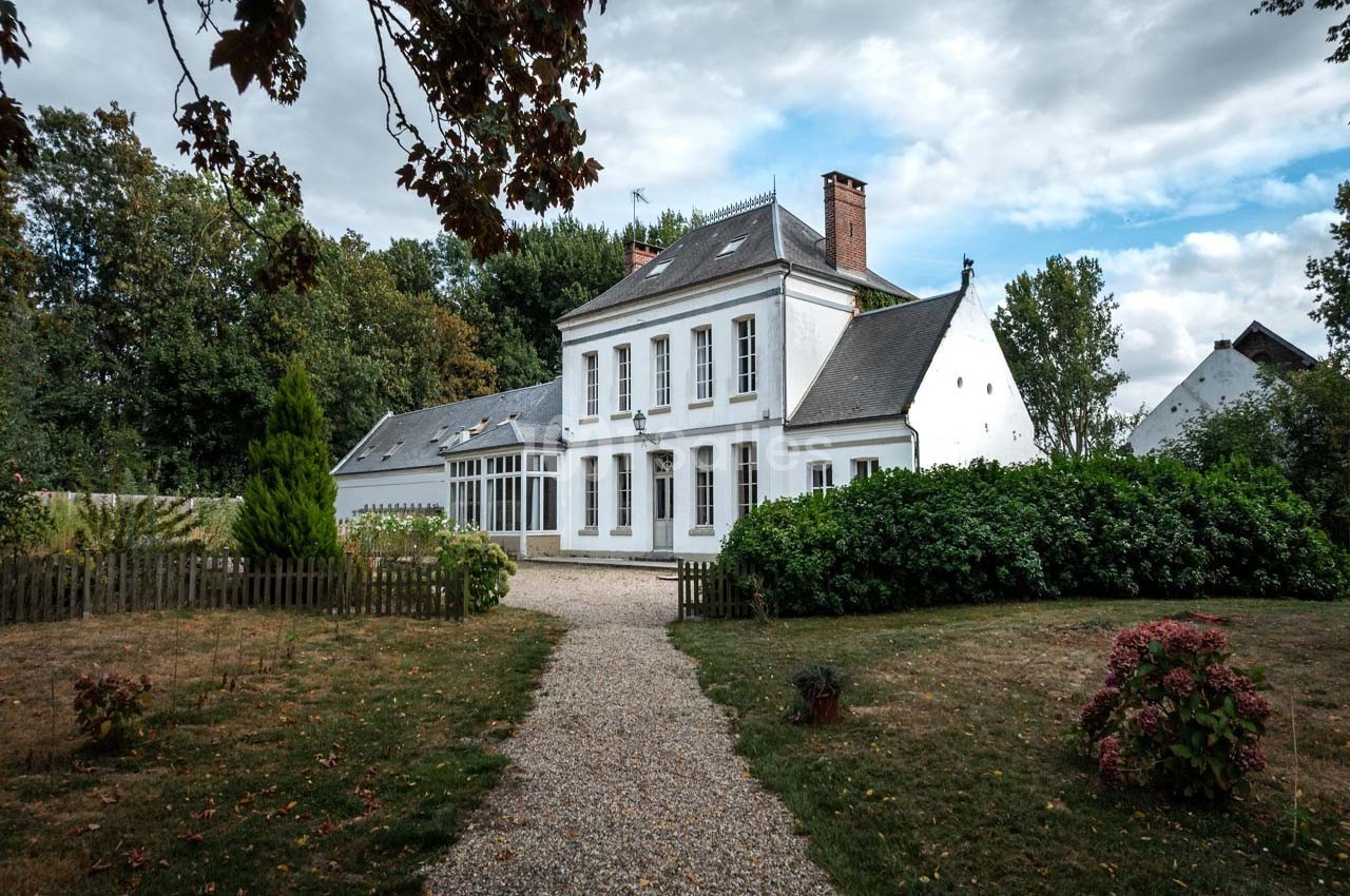 Grande maison blanche à deux étages avec jardin, allée gravillonnée et arbres en arrière-plan sous un ciel nuageux.