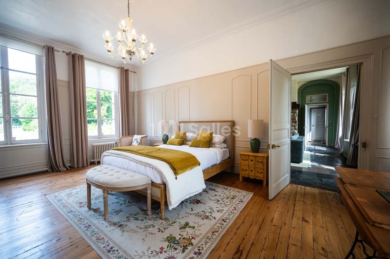 Chambre lumineuse avec lit double, couvre-lit jaune, tapis floral, parquet en bois et grandes fenêtres avec rideaux.