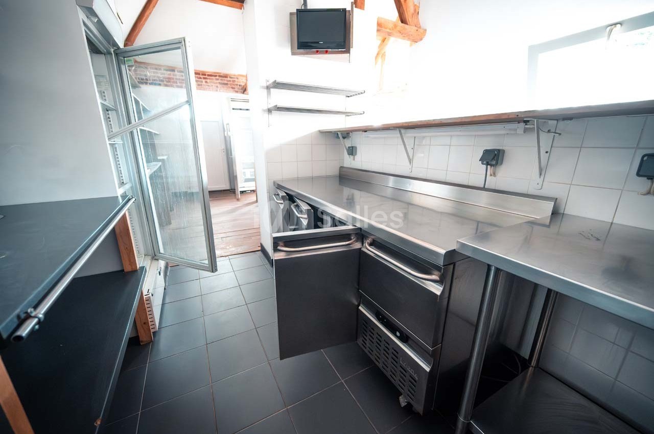 Cuisine professionnelle avec plans de travail en inox, étagères murales, réfrigérateur ouvert et carrelage sombre.