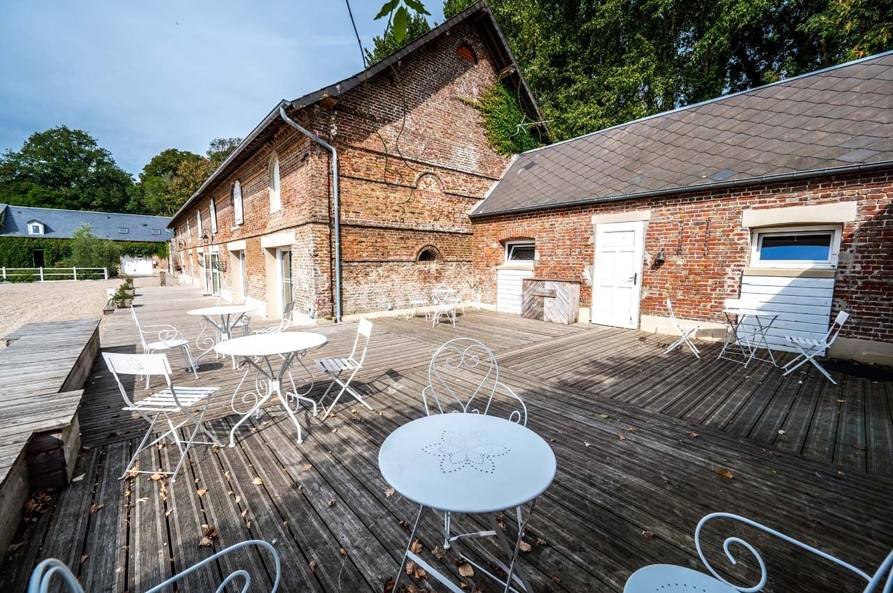 Terrasse en bois avec tables et chaises blanches devant un bâtiment en briques, entourée de verdure.