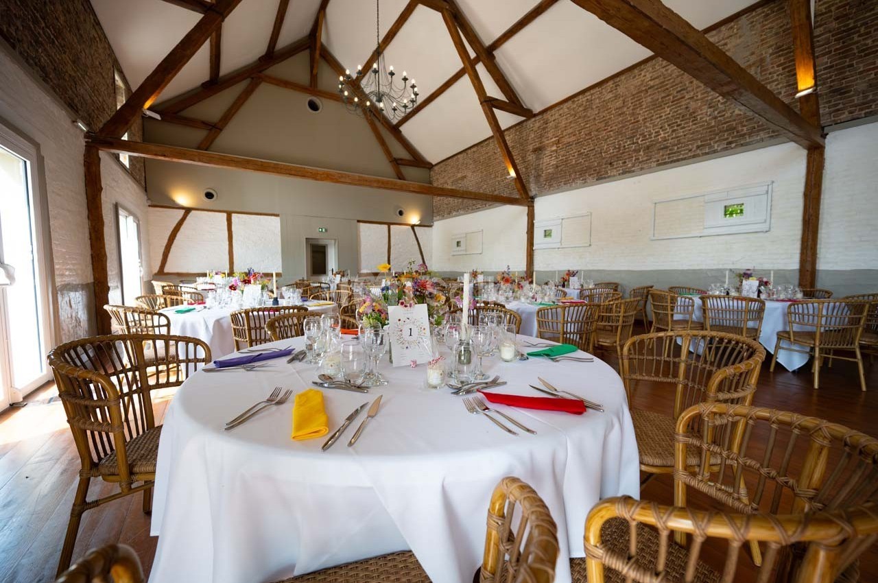 Salle de réception lumineuse avec tables rondes dressées, chaises en rotin et poutres apparentes au plafond.