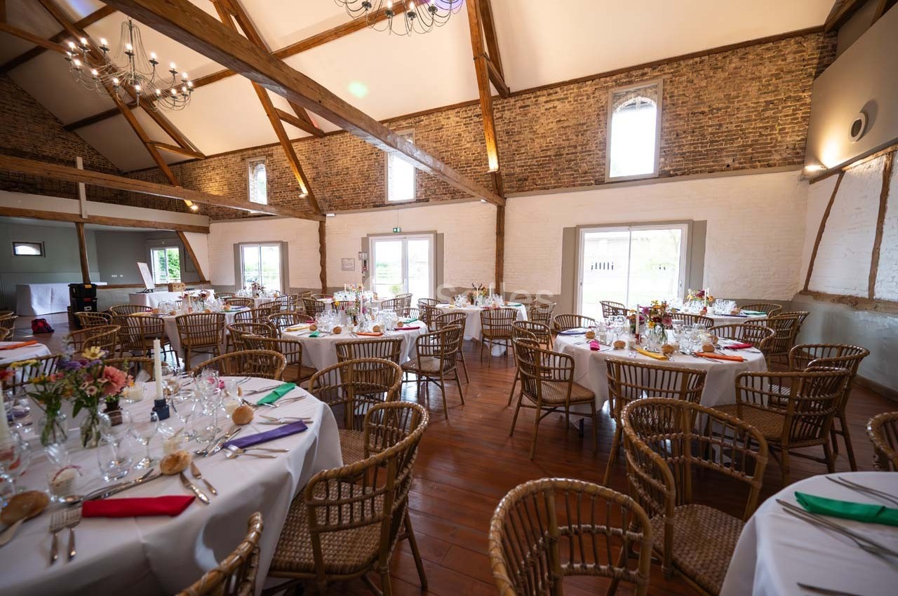 Salle de réception lumineuse avec murs en briques, poutres apparentes et tables rondes décorées pour un événement.