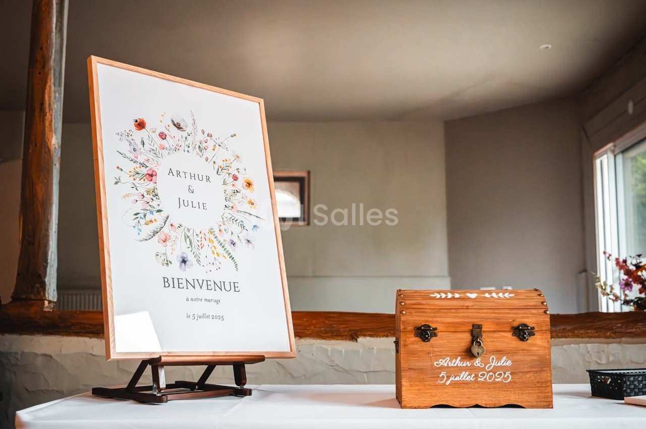 Panneau de bienvenue à un mariage avec des fleurs illustrées, posé sur un chevalet, à côté d'une boîte en bois gravée.