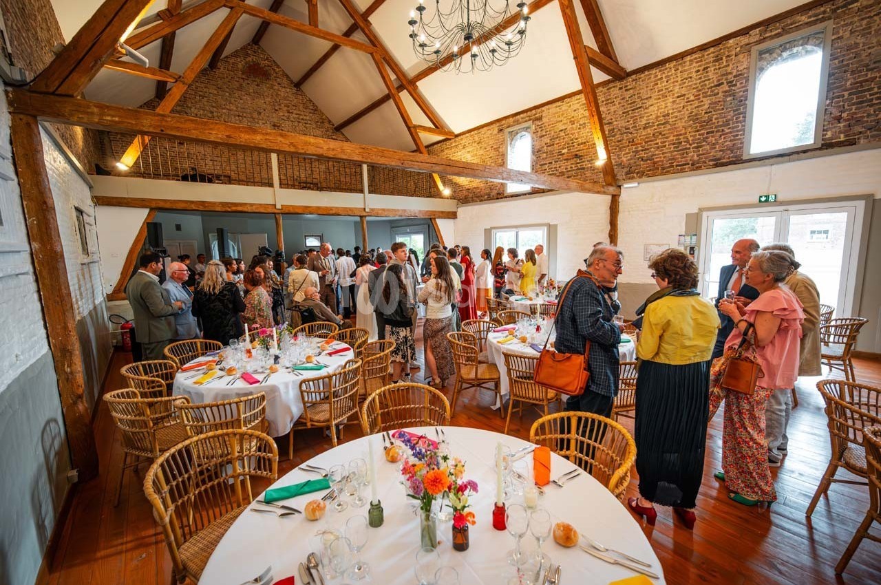 Salle de réception avec des invités debout et assis autour de tables décorées, sous une charpente en bois apparente.