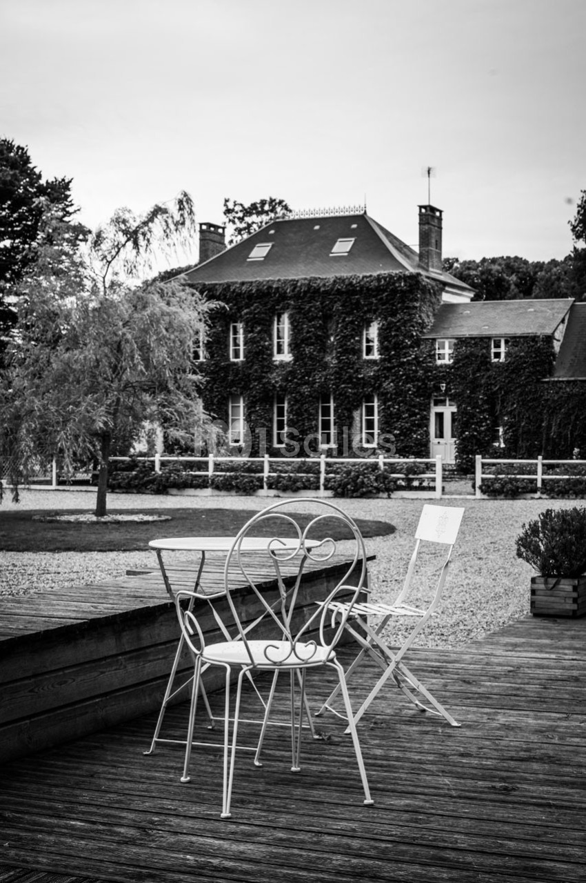 Chaises et table en fer forgé sur une terrasse en bois, avec une maison couverte de lierre en arrière-plan.