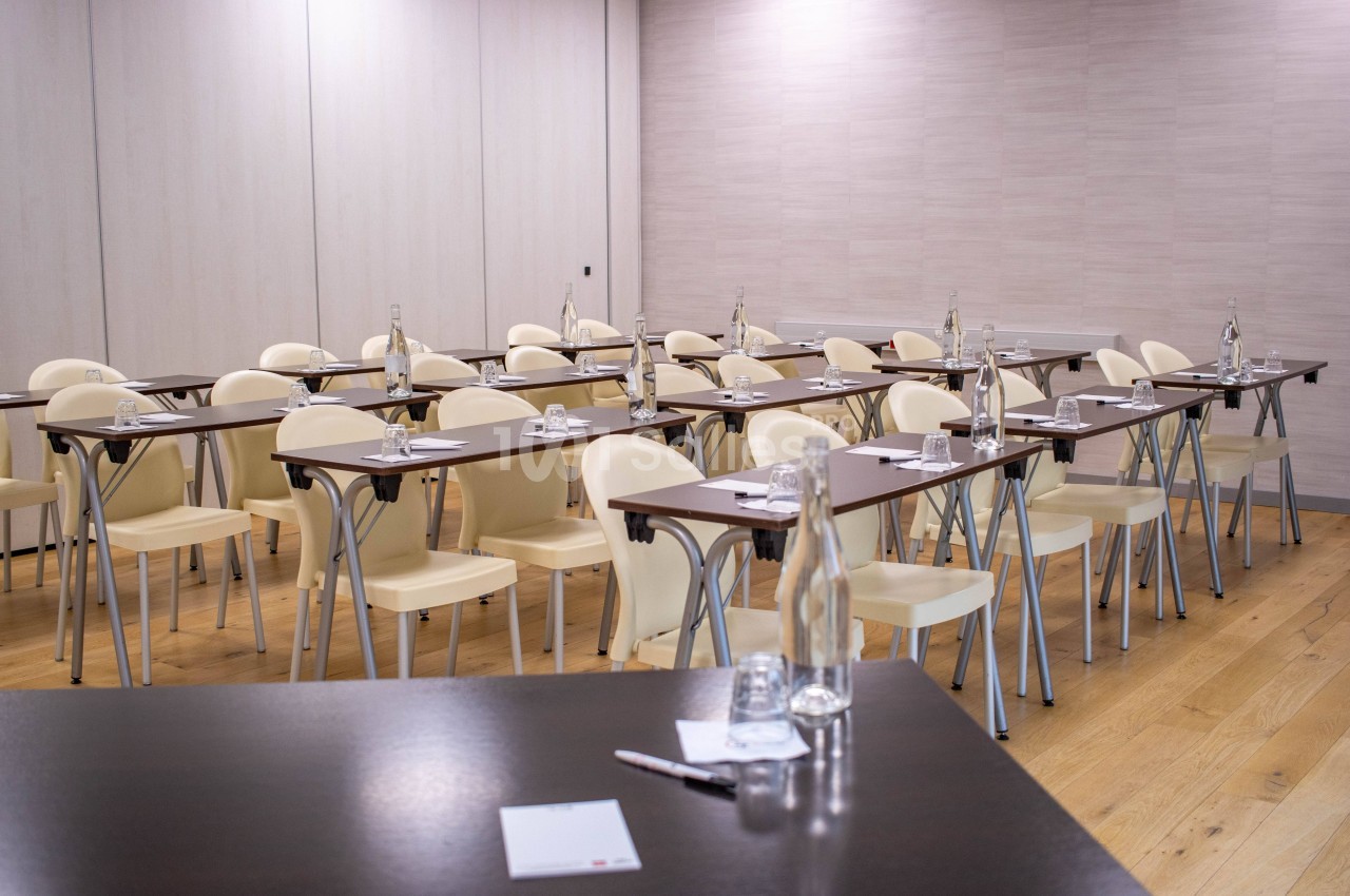 Salle de réunion avec des tables et chaises alignées, disposées en format classe, avec bouteilles et verres sur les tables.