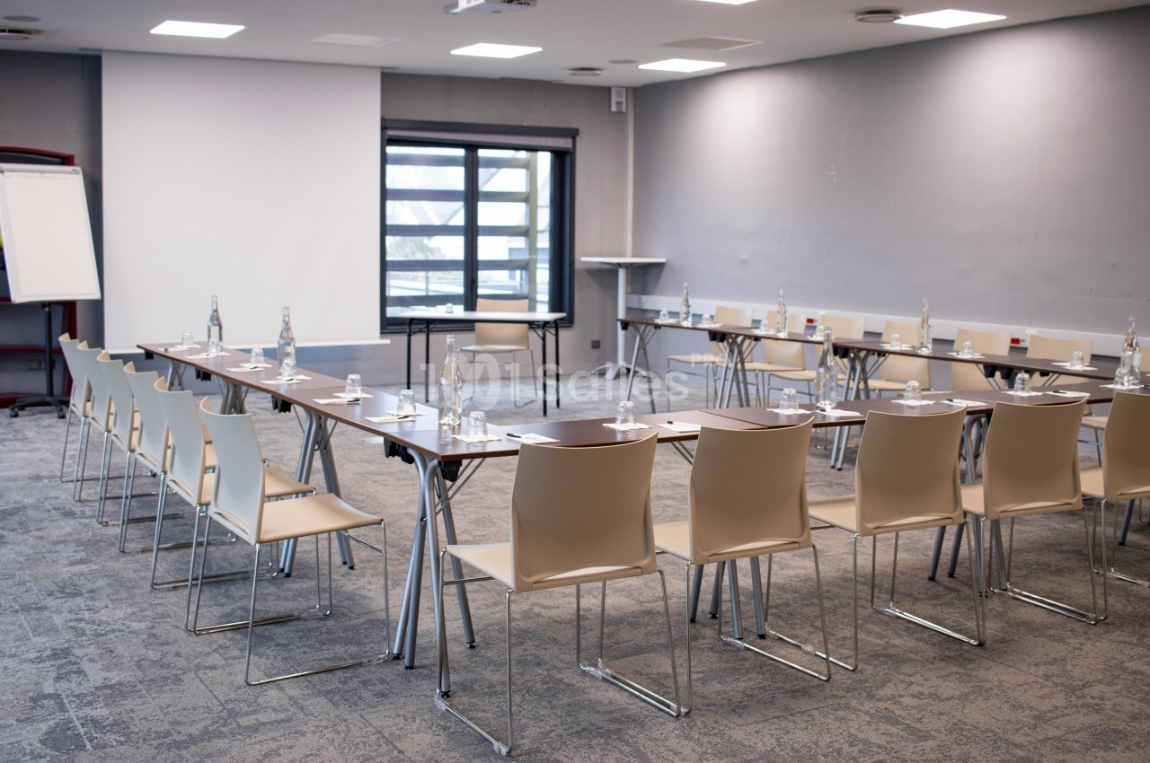 Salle de réunion lumineuse avec tables disposées en U, chaises, bouteilles d'eau et matériel de présentation.