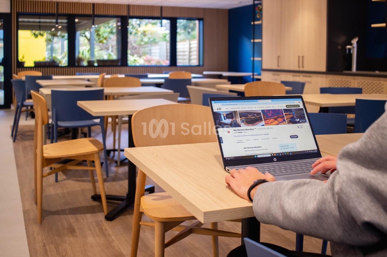 Une personne utilise un ordinateur portable dans une salle lumineuse avec des tables et des chaises en bois et en métal.