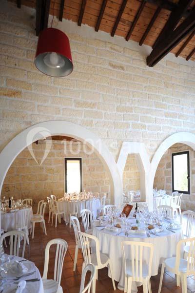 Salle lumineuse avec murs en pierre, tables rondes dressées avec nappes blanches et chaises blanches, plafond en bois.