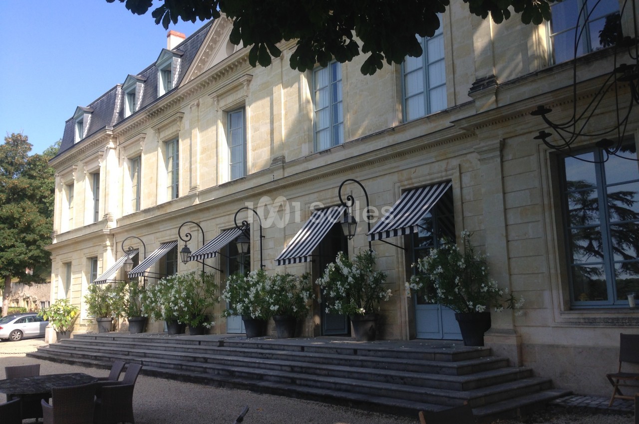 Façade d'un bâtiment en pierre avec des auvents rayés, des pots de fleurs et une terrasse ombragée.