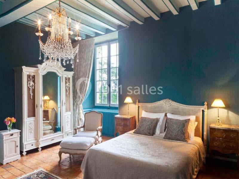 Chambre élégante avec murs bleus, lit double, armoire blanche, lustre en cristal et grande fenêtre laissant entrer la…