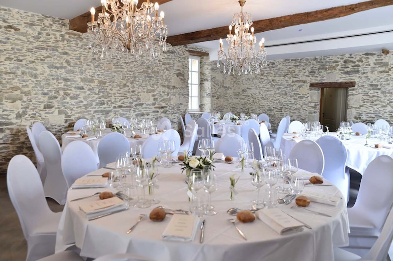 Salle de réception avec murs en pierre, tables rondes dressées avec nappes blanches, chandeliers et décoration élégante.