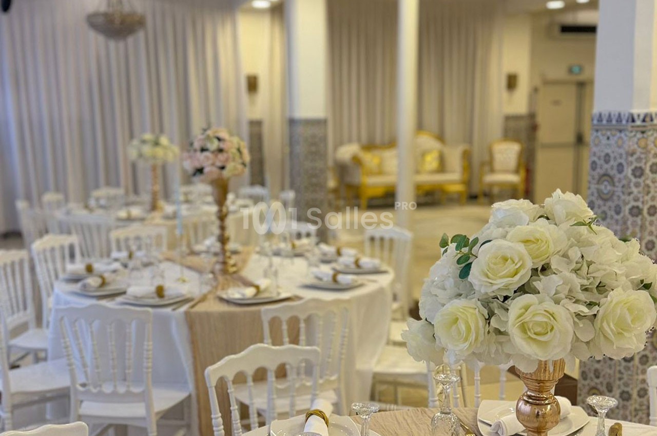 Salle de réception décorée avec des tables rondes, nappes blanches, chaises assorties et centres de table fleuris.
