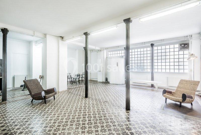 Espace lumineux avec sol en carreaux de ciment, colonnes métalliques, chaises et fauteuils en osier.