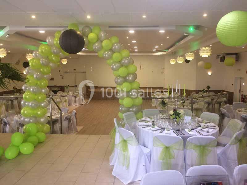 Salle de réception décorée avec des ballons verts et blancs, tables dressées et arche de ballons au centre.