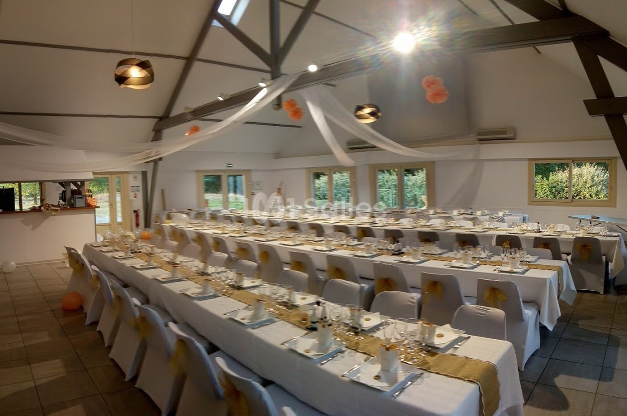 Salle de réception décorée avec des tables alignées, nappes blanches, chaises habillées et guirlandes suspendues.