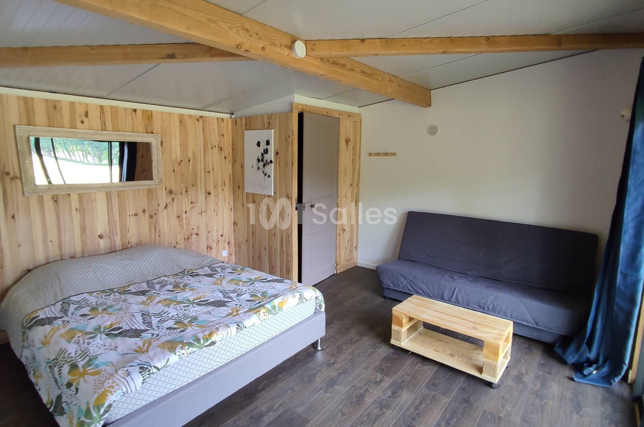 Chambre lumineuse avec lit double, canapé, table basse en bois, murs en bois clair et poutres apparentes.