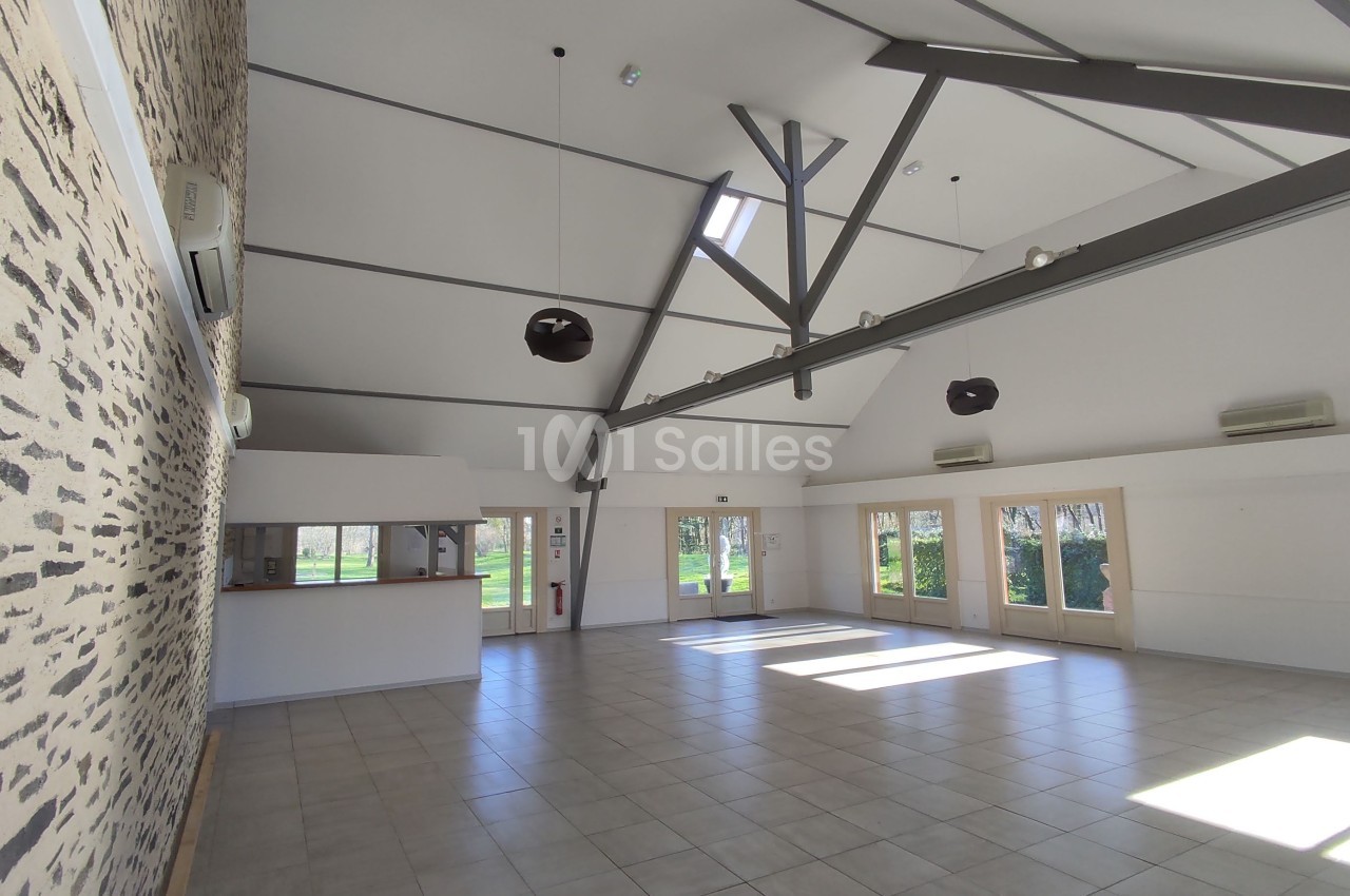 Salle lumineuse avec plafond haut, poutres apparentes, murs en pierre et grandes baies vitrées donnant sur un jardin.