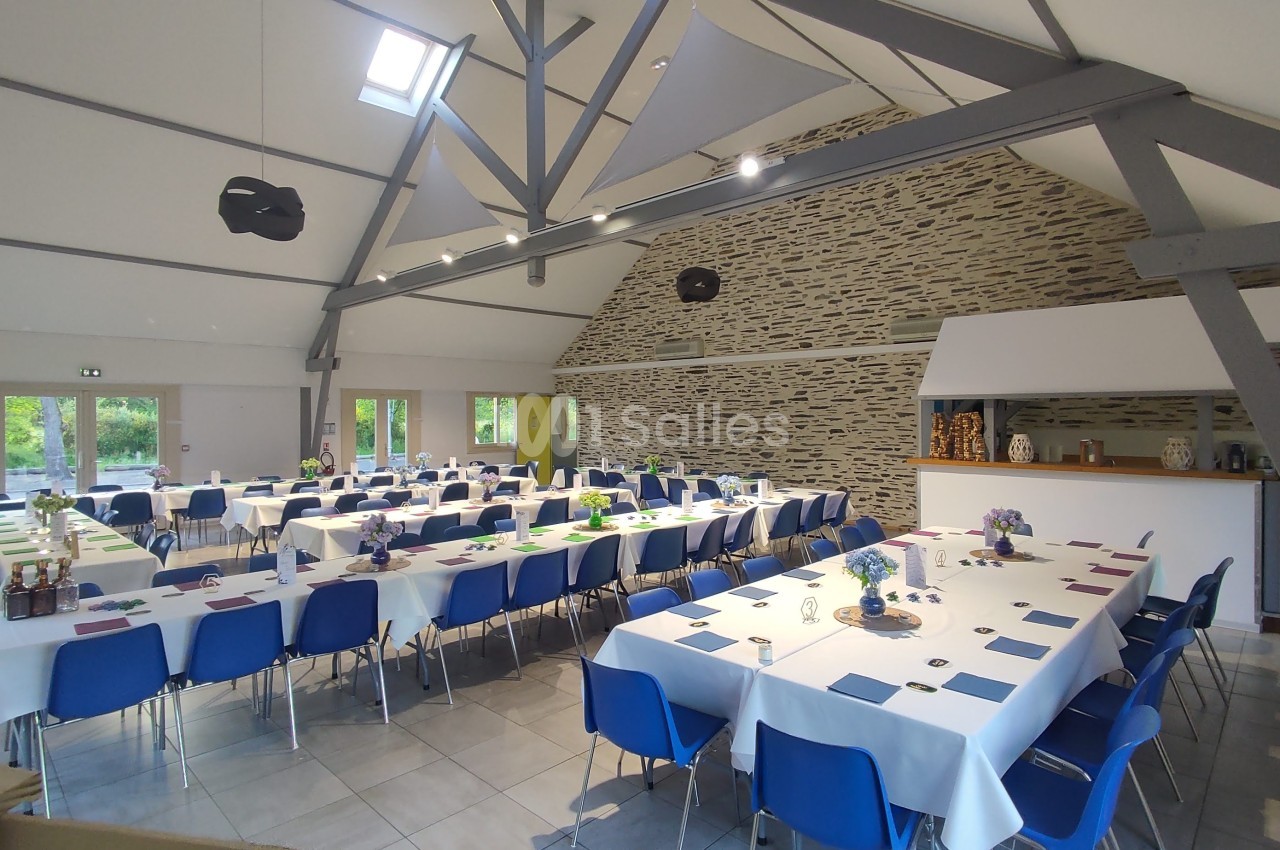 Salle lumineuse avec tables dressées en U, nappes blanches, chaises bleues et murs en pierre apparente.
