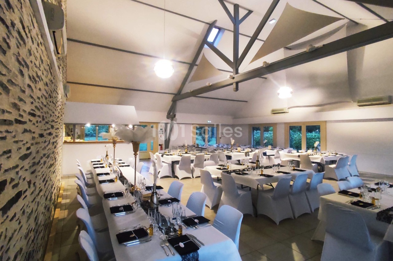 Salle de réception décorée avec des tables alignées, nappes blanches et chaises houssées, éclairée par des suspensions.