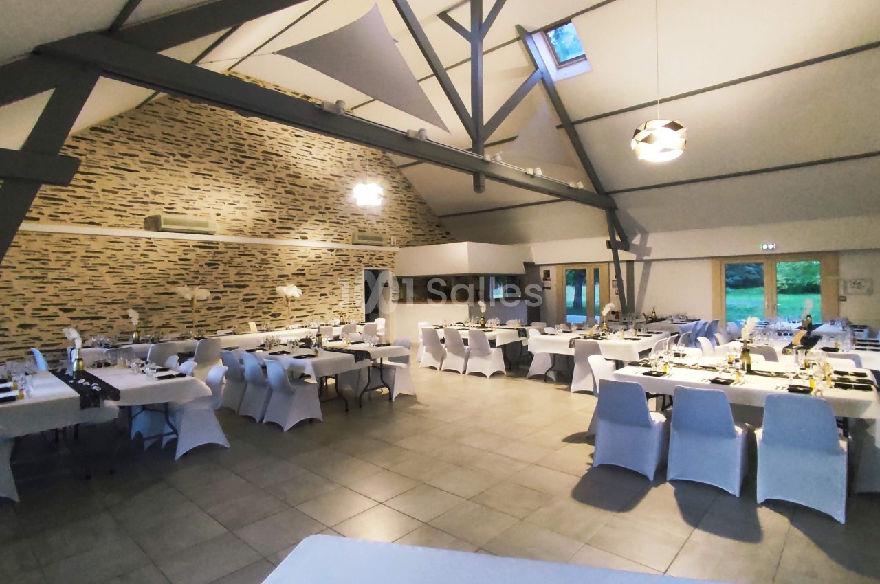 Salle de réception spacieuse avec tables dressées, chaises blanches et murs en pierre apparente.