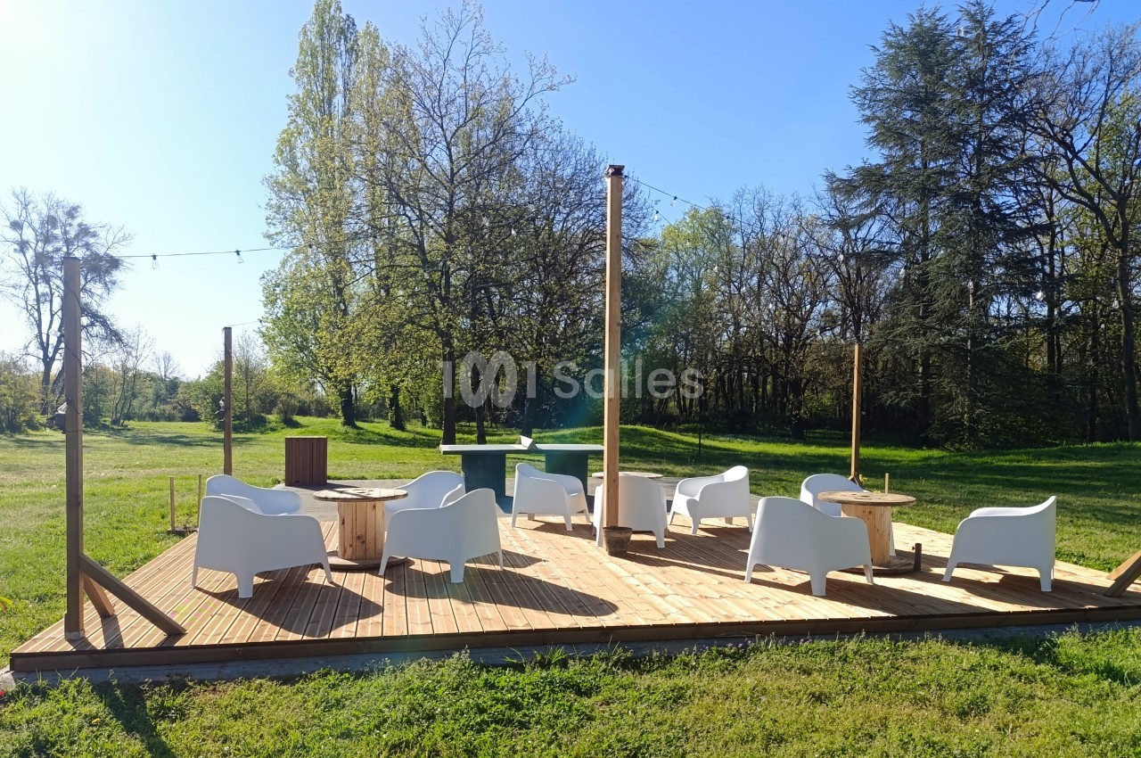 Terrasse en bois avec fauteuils blancs et tables basses, située dans un espace vert entouré d'arbres.
