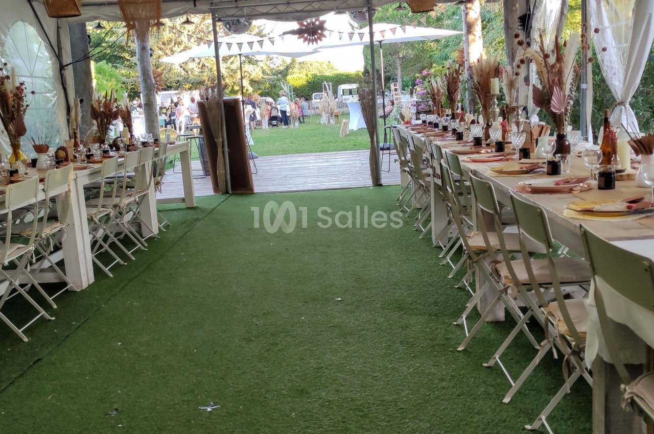 Tables décorées pour un événement sous une tente, avec chaises blanches et allée centrale sur gazon artificiel.