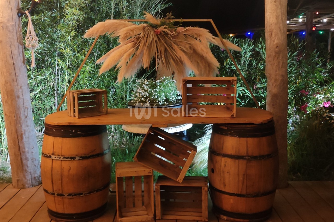 Deux tonneaux en bois soutenant une planche décorée de caisses en bois, de fleurs et de pampas, en extérieur nocturne.