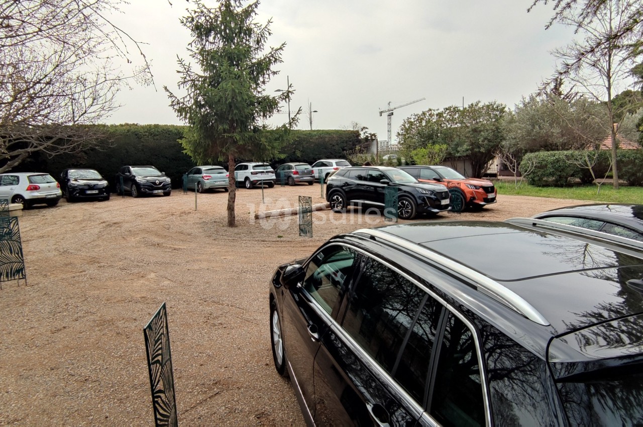 Parking extérieur avec plusieurs voitures stationnées sur un sol en gravier, entouré d'arbres et de verdure.