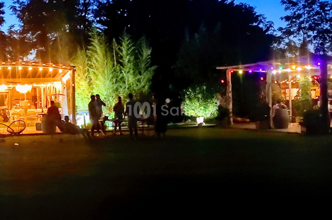 Deux terrasses éclairées par des guirlandes lumineuses dans un jardin en soirée, avec des personnes rassemblées.