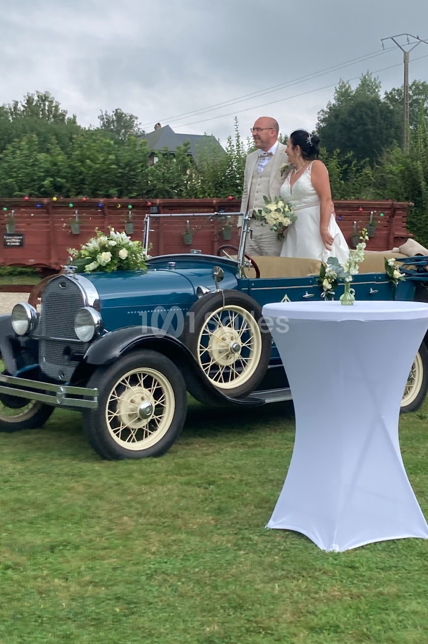 Un couple en tenue de mariage pose près d'une voiture ancienne bleue décorée de fleurs, sur une pelouse.