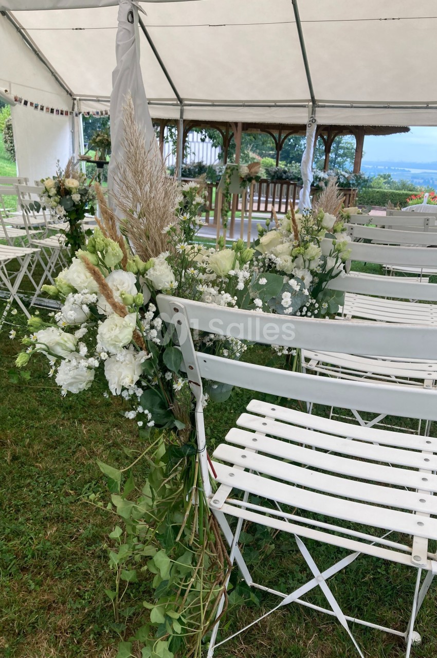 Chaises blanches pliantes décorées de fleurs blanches et feuillages sous une tente, dans un cadre extérieur verdoyant.