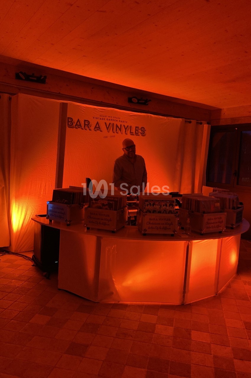 Un DJ mixe derrière un comptoir éclairé en orange, entouré de vinyles, dans une ambiance tamisée.