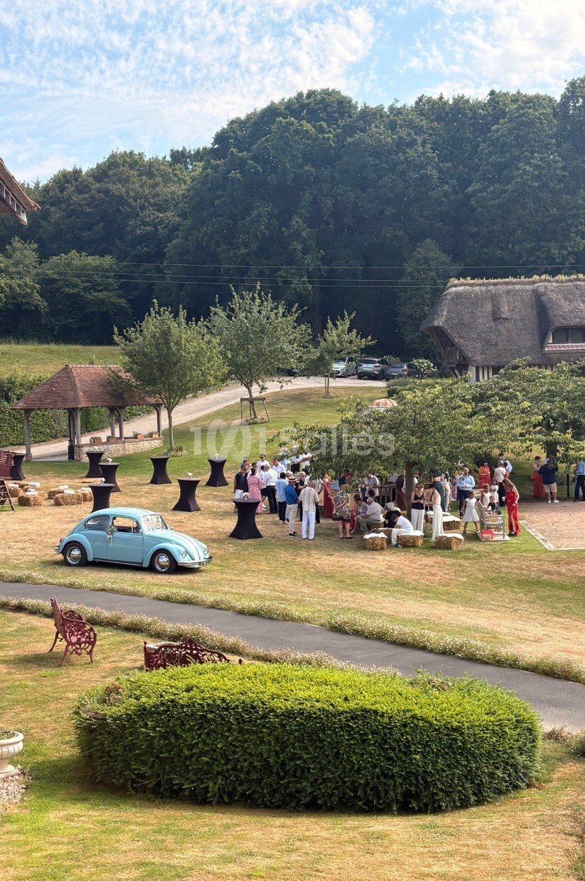 Groupe de personnes rassemblées en extérieur près de tables hautes, avec une voiture bleue et un paysage champêtre en…