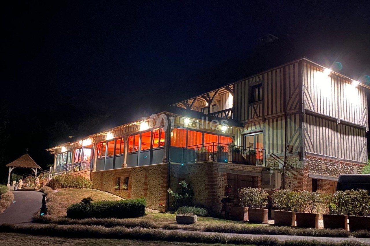 Bâtiment à colombages éclairé la nuit, avec une terrasse vitrée illuminée et un jardin environnant.