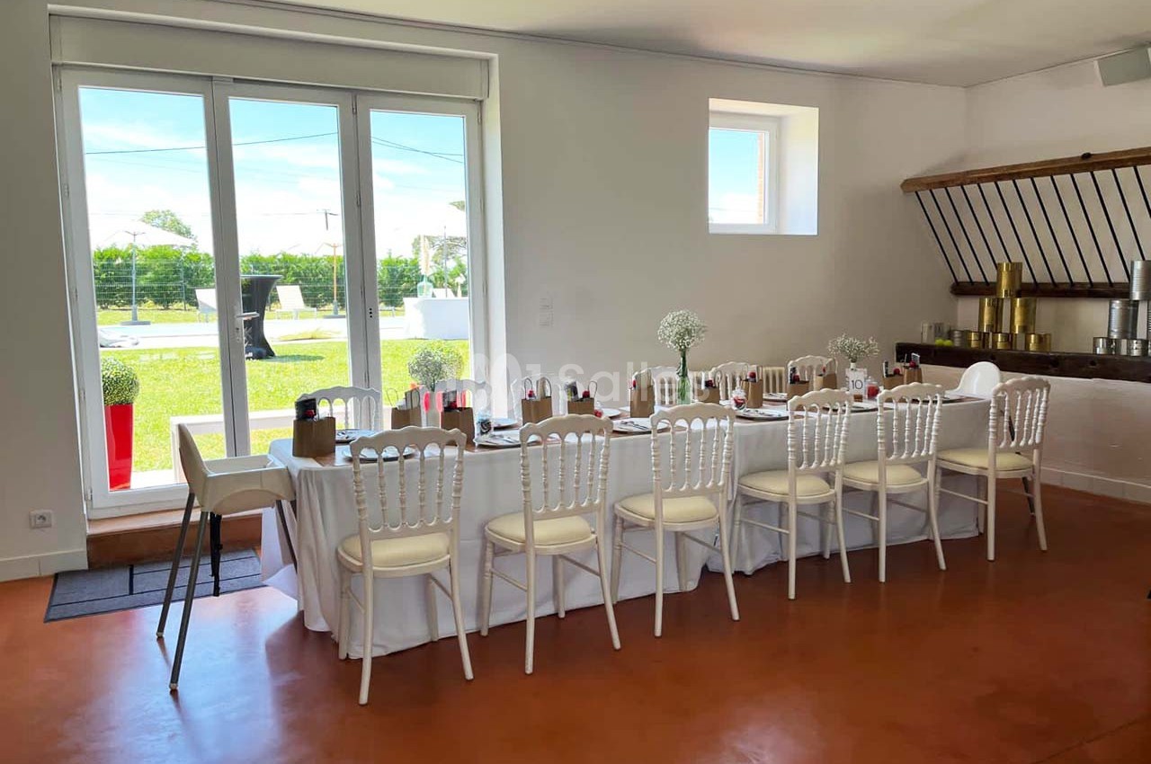 Salle lumineuse avec une grande table dressée pour un repas, chaises blanches et vue sur un jardin à travers une baie vitrée.