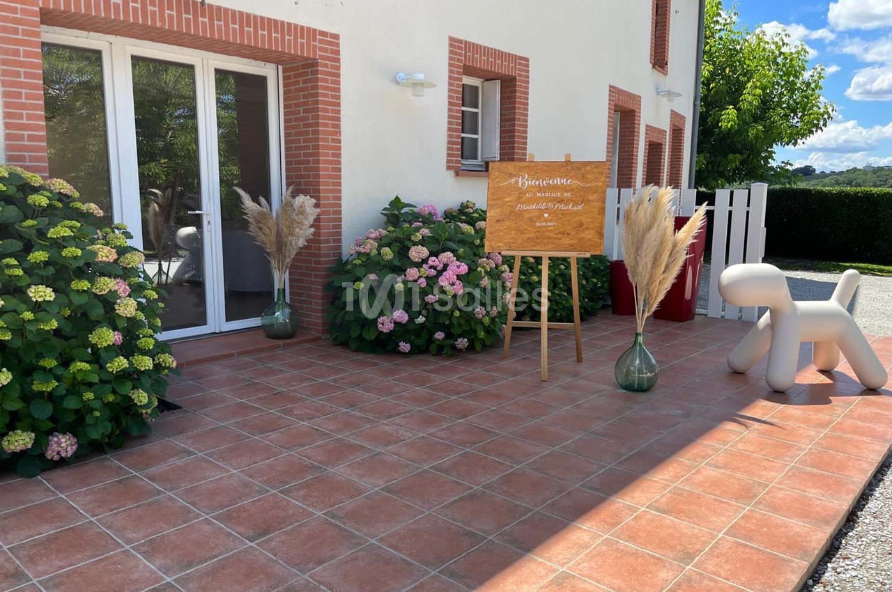 Terrasse carrelée avec plantes, panneau de bienvenue sur chevalet, vases décoratifs et siège en forme de chien.