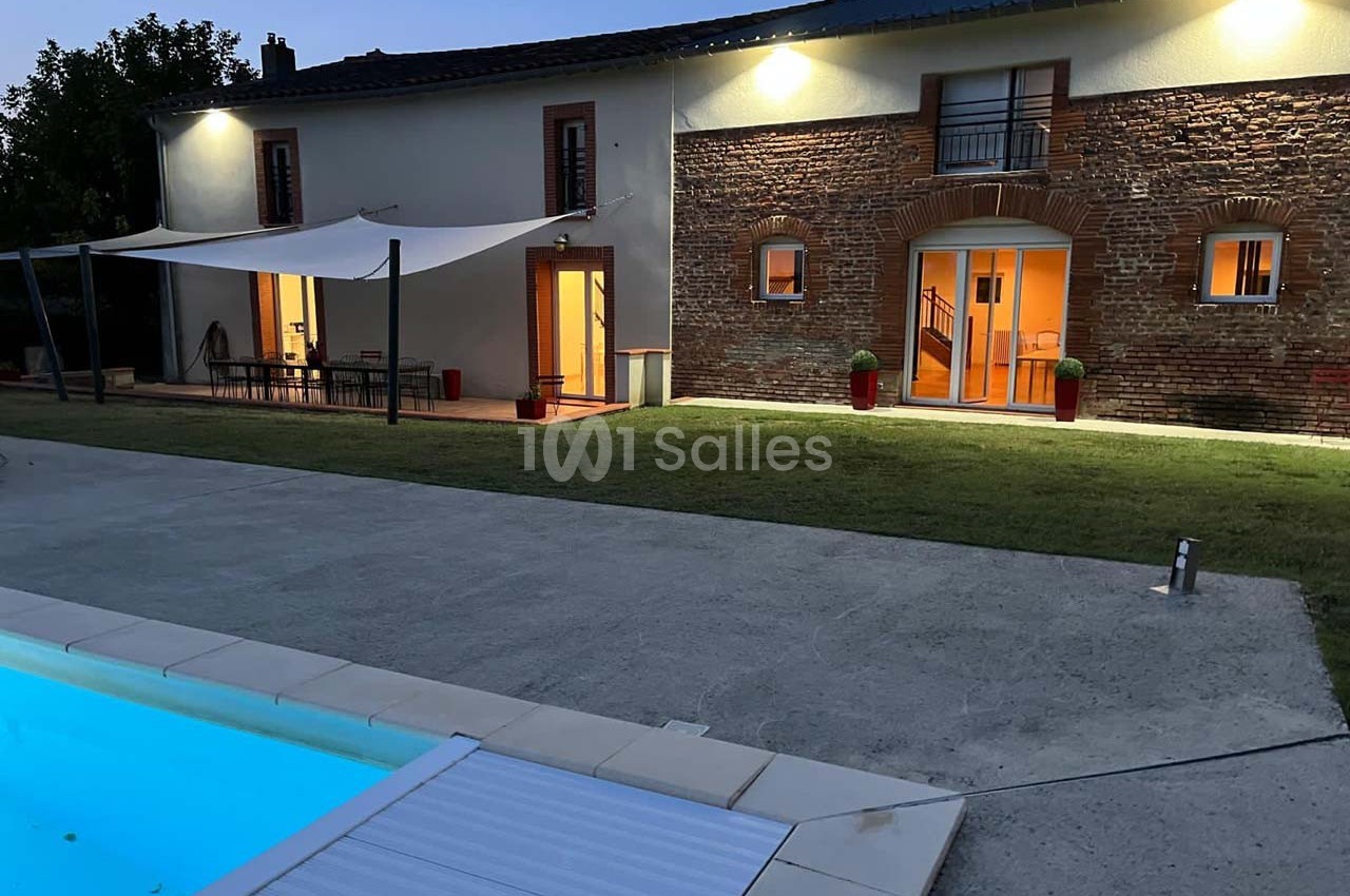 Maison en briques éclairée à la tombée de la nuit, avec terrasse couverte, jardin et piscine au premier plan.