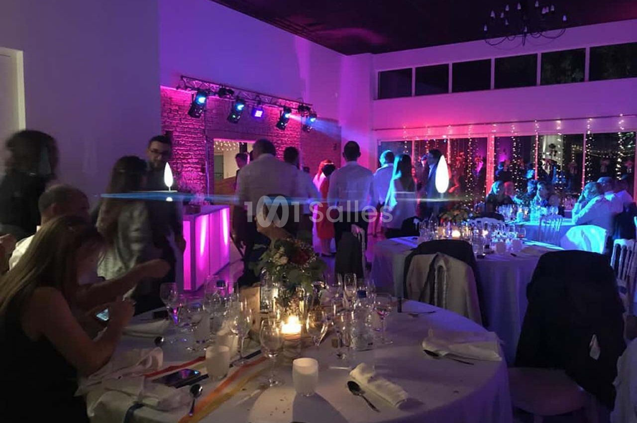 Salle de réception éclairée avec des lumières colorées, des invités debout et des tables dressées pour un événement.