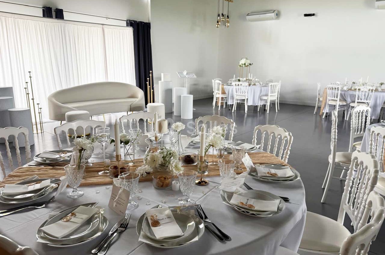 Salle de réception lumineuse avec tables rondes dressées, nappes blanches, décorations florales et chaises élégantes.
