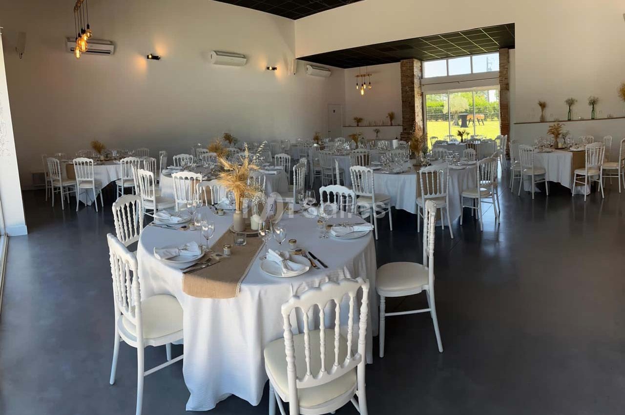 Salle de réception lumineuse avec tables rondes dressées, nappes blanches et décorations florales naturelles.