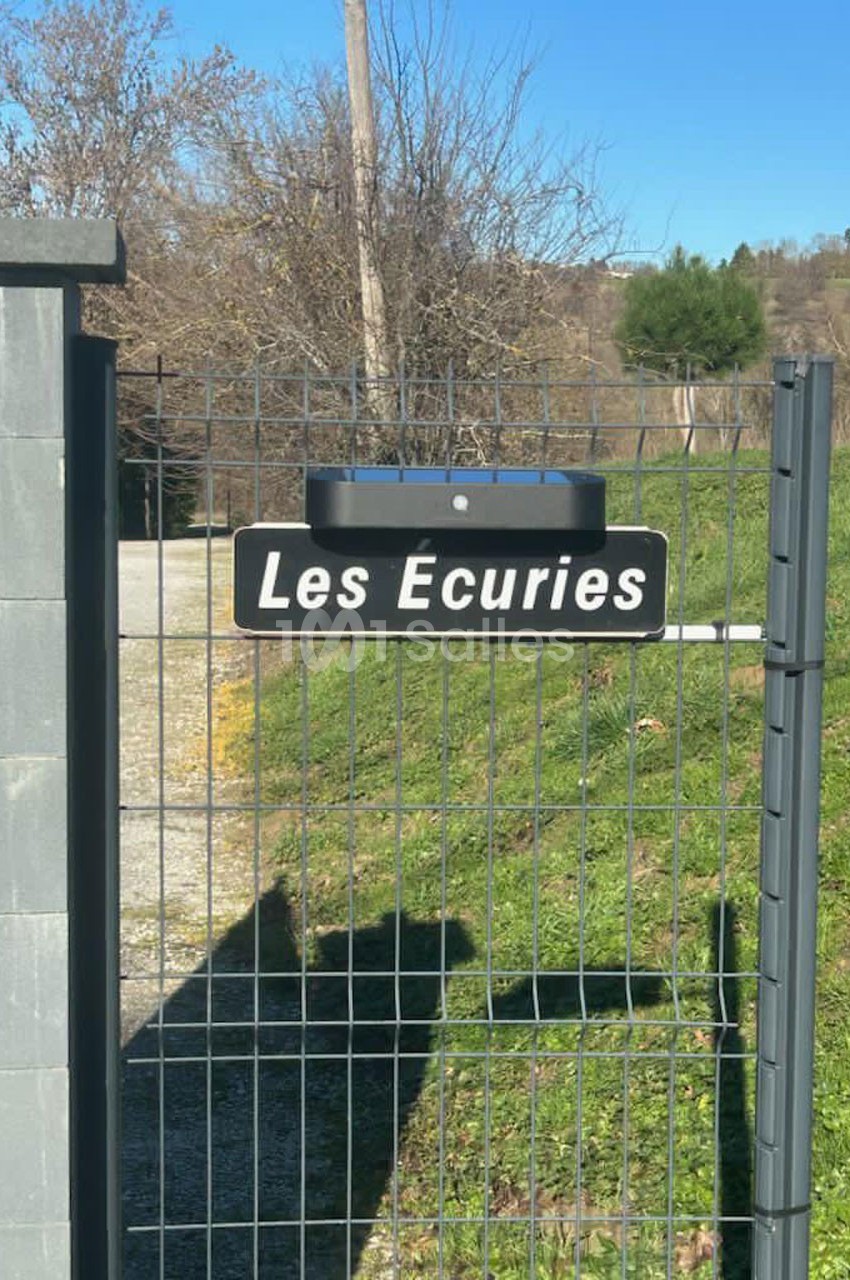 Panneau ’Les Écuries’ fixé sur un portail grillagé, avec un chemin et des arbres en arrière-plan.