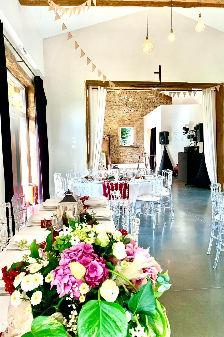 Salle de réception lumineuse décorée pour un événement, avec tables dressées, fleurs et guirlandes suspendues.
