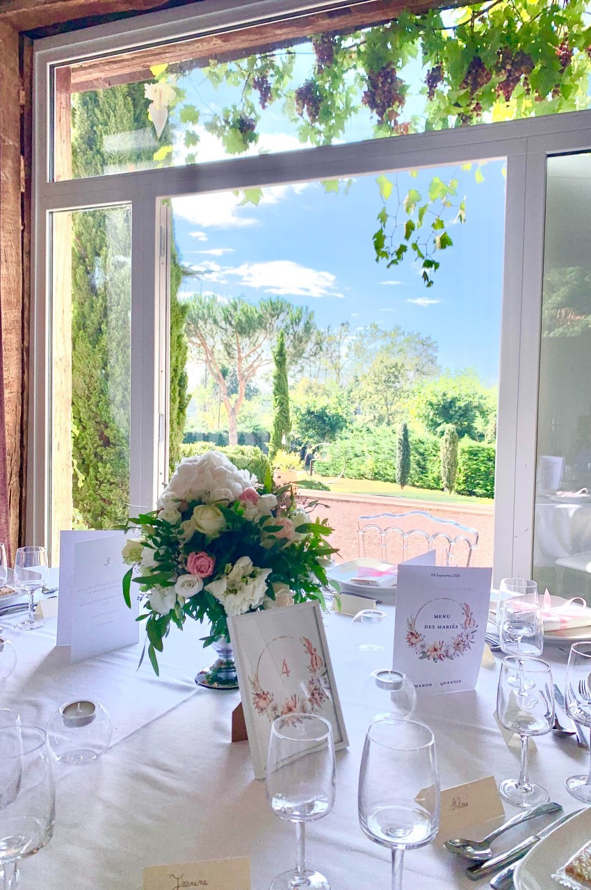 Table élégamment dressée avec des fleurs et des menus, donnant sur un jardin verdoyant visible à travers une baie vitrée.
