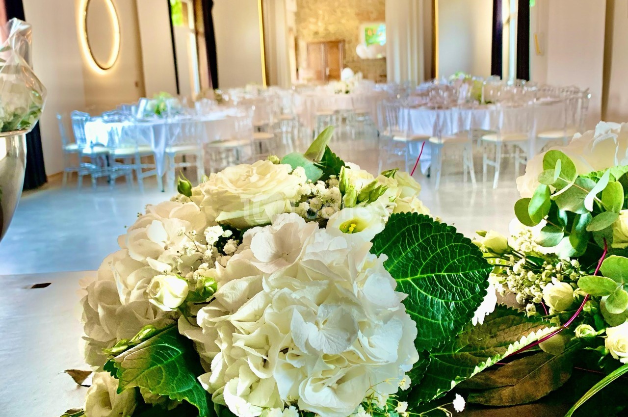Salle de réception lumineuse décorée pour un événement, avec des bouquets de fleurs blanches au premier plan.