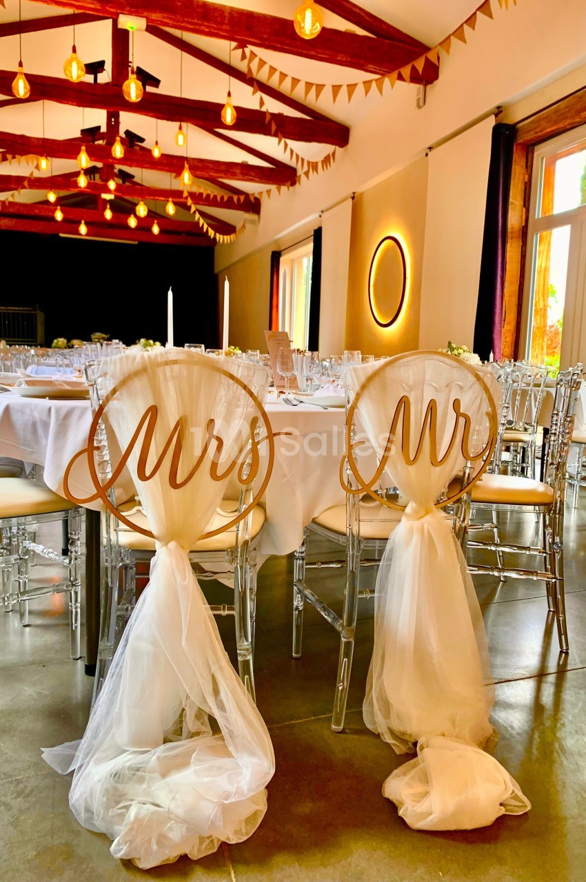 Chaises décorées avec les inscriptions ’Mrs’ et ’Mr’, entourées de tables dressées dans une salle de réception lumineuse.