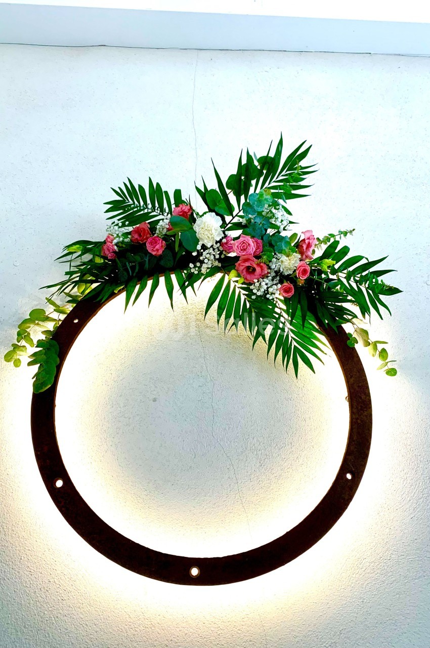 Couronne lumineuse circulaire décorée de feuillage vert et de fleurs roses et blanches, accrochée à un mur clair.